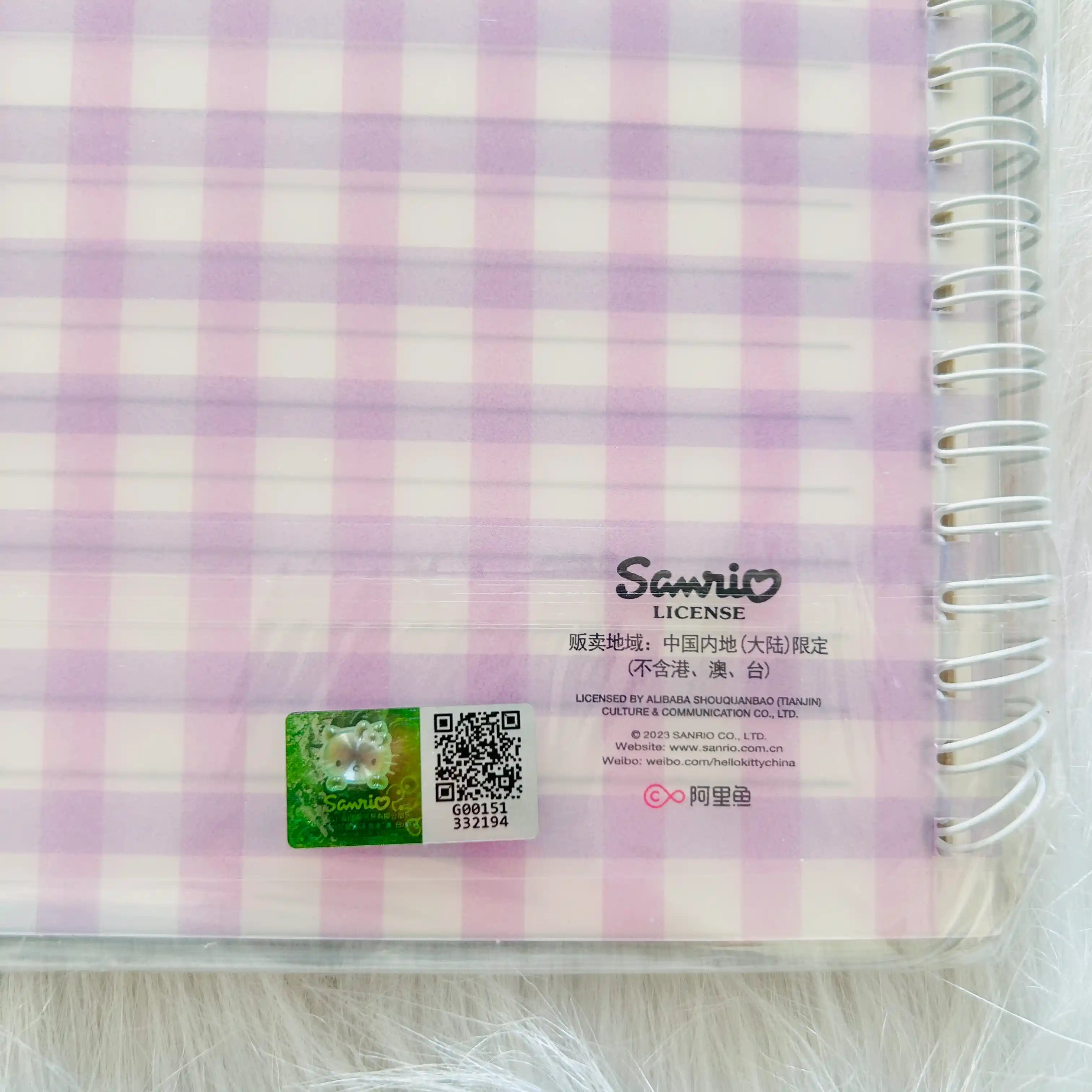 A close-up of a spiral-bound notebook with a pink and white gingham patterned cover, featuring a Sanrio License sticker.