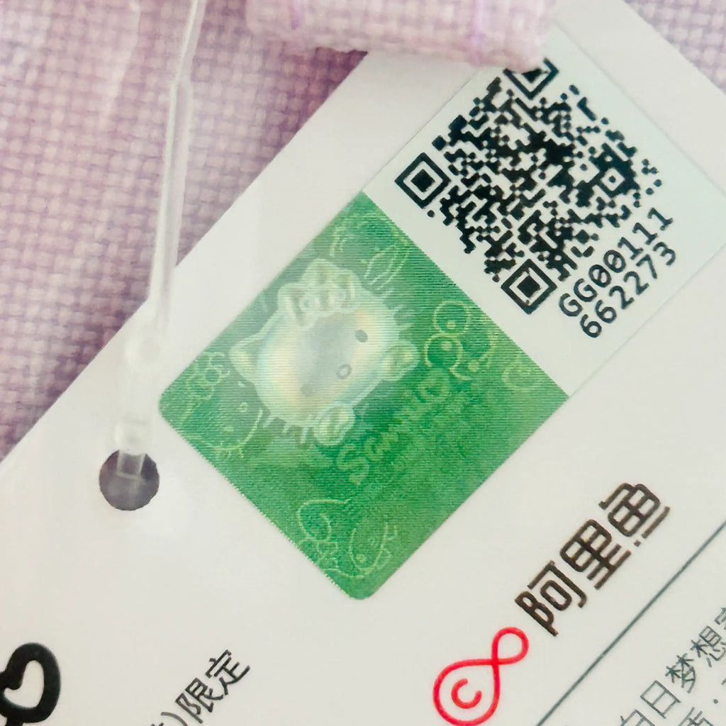 A close-up of a white garment tag attached to a light pink textured fabric. The tag features a green holographic Hello Kitty-style character sticker, a black QR code, and Chinese text, including the AliExpress logo.