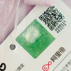 A close-up of a white garment tag attached to a light pink textured fabric. The tag features a green holographic Hello Kitty-style character sticker, a black QR code, and Chinese text, including the AliExpress logo.