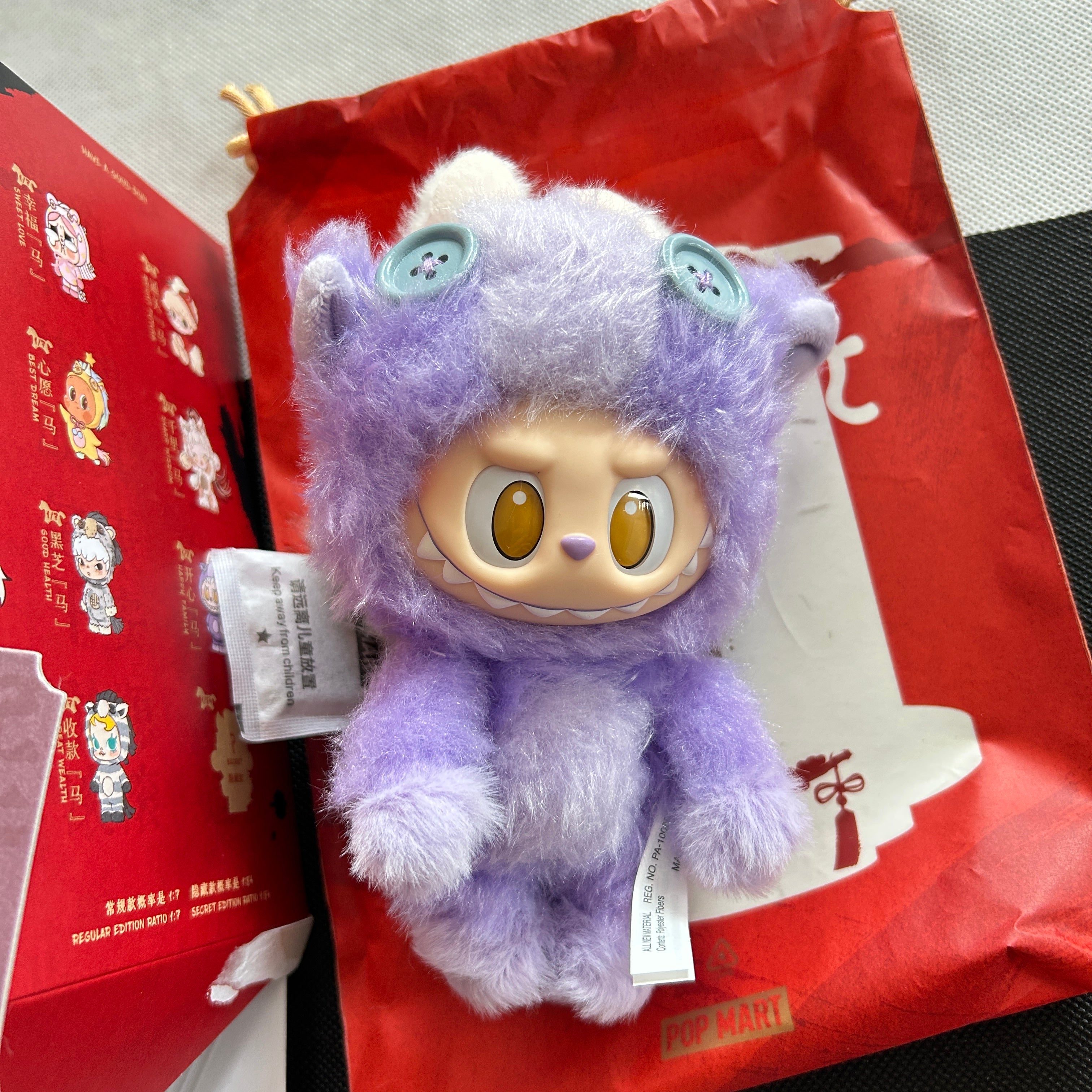 A purple fuzzy Labubu Have a Good Rub plush toy, placed on top of a red printed box and bag.  