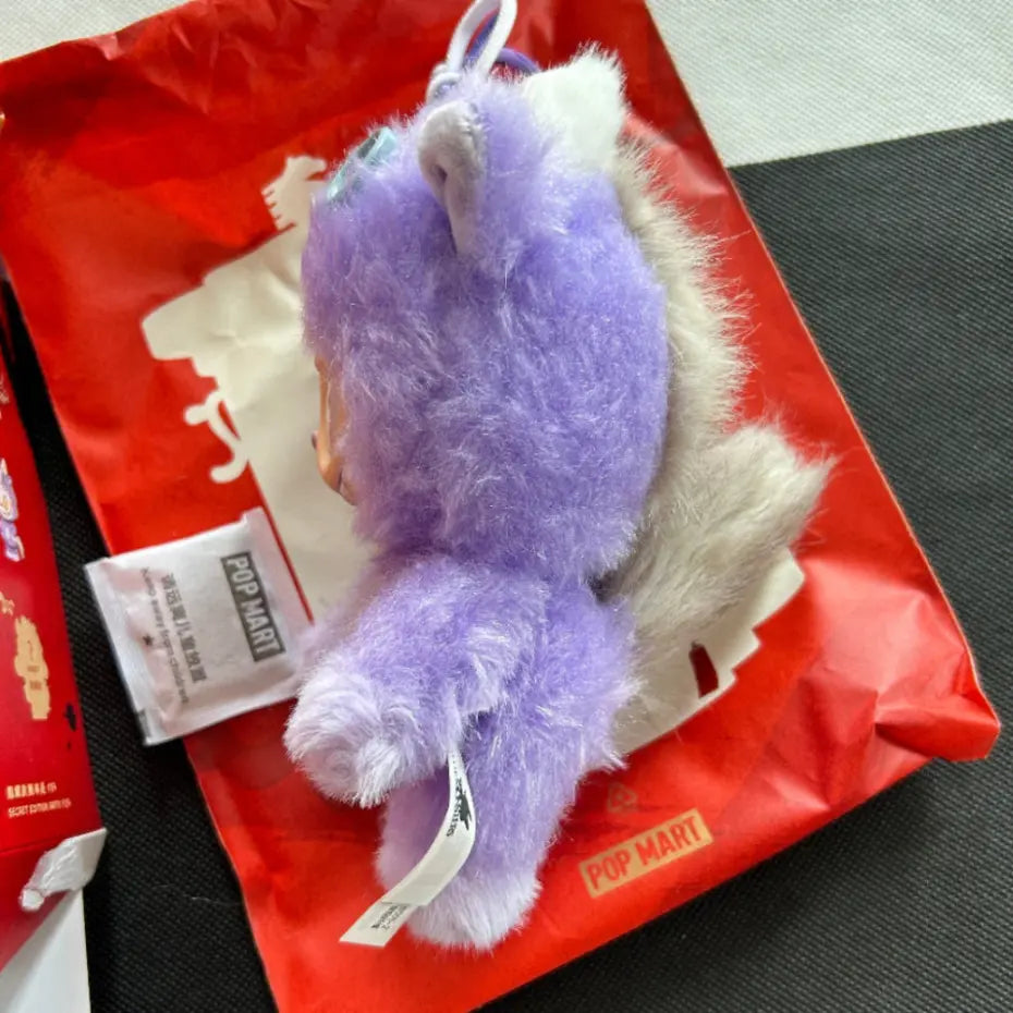 A purple Labubu Happy Family plush pendant, placed on top of a red POP MART shopping bag with a product tag attached.