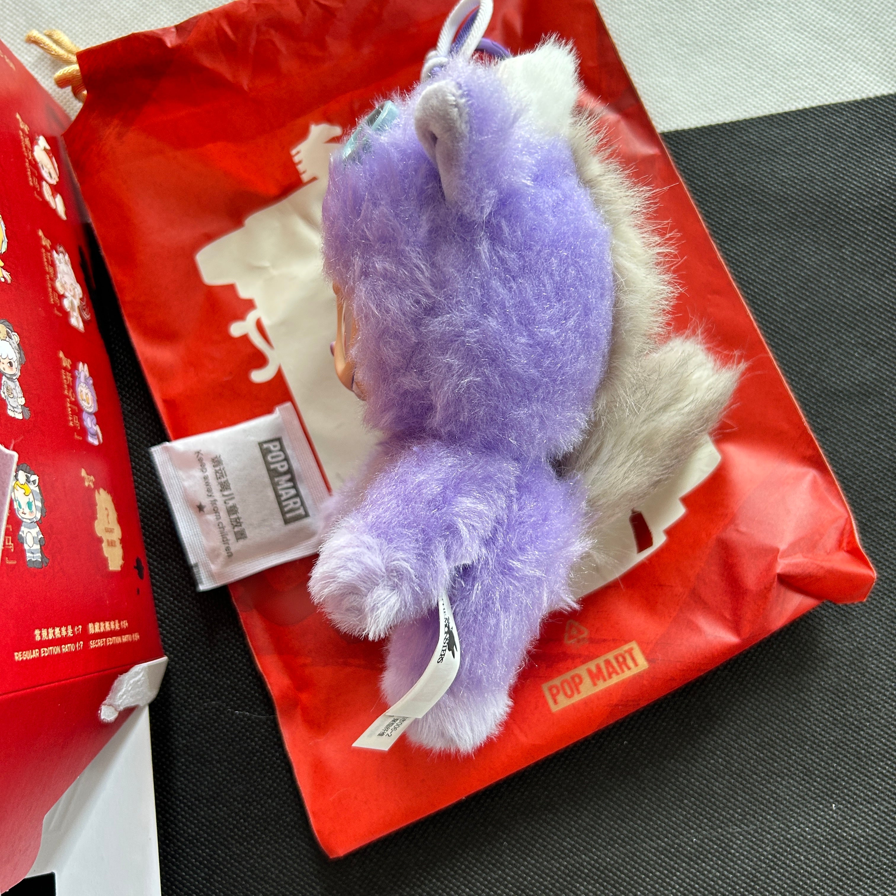A purple Labubu Happy Family plush pendant, placed on top of a red POP MART shopping bag with a product tag attached.