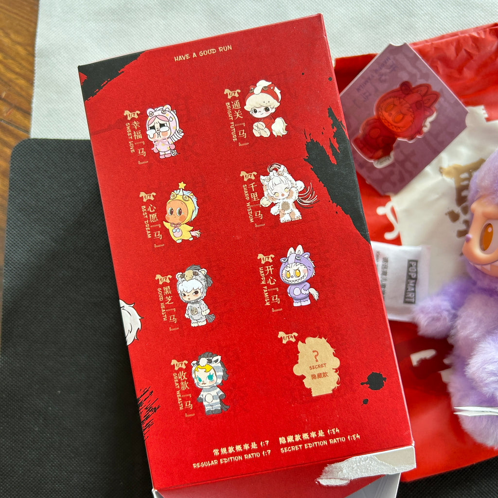 A red blind box featuring cartoon character illustrations on the front, photographed alongside other colorful packaging and plush items on a tabletop.