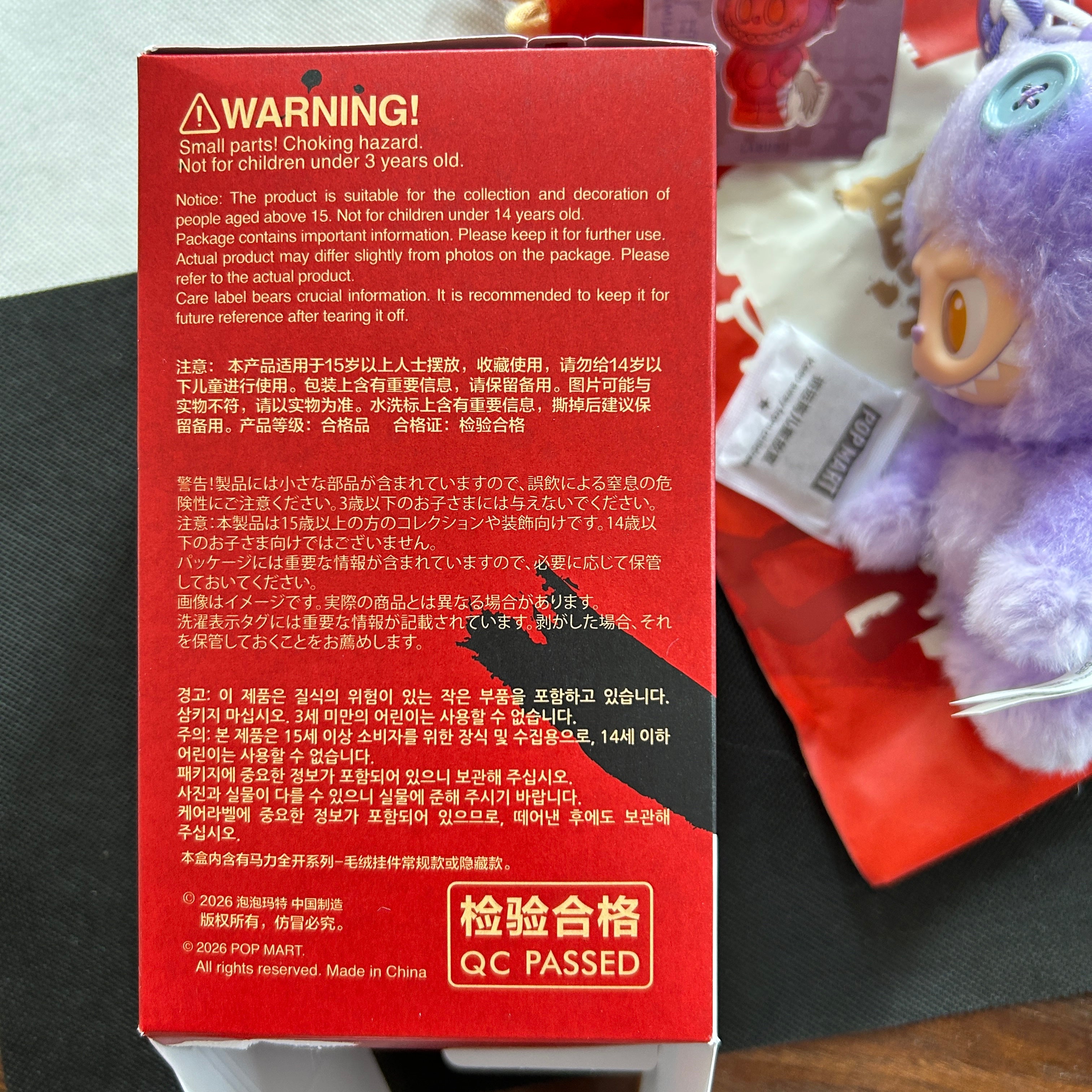 A red cardboard box with Chinese warning text and a yellow 'QC PASSED' label, photographed on a dark surface next to a small plush toy.