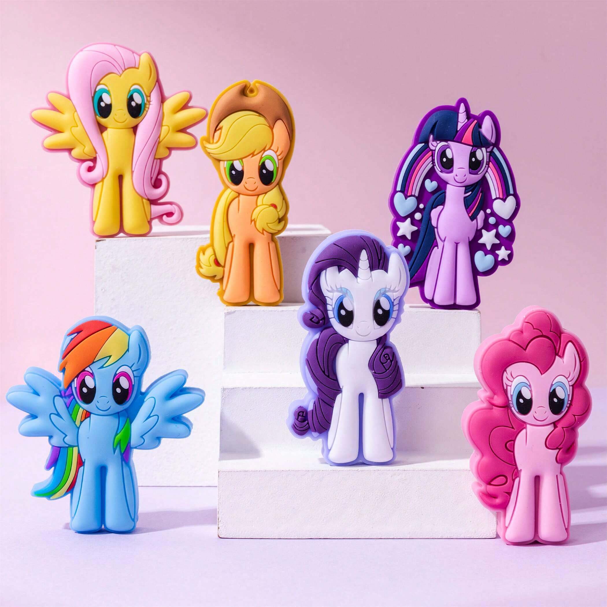 Six My Little Pony character figurines displayed on white step risers against a soft pink background. The characters include Flory Meow, Applejack, Twilight Sparkle, Rainbow Dash, Rarity, and Pinkie Pie.