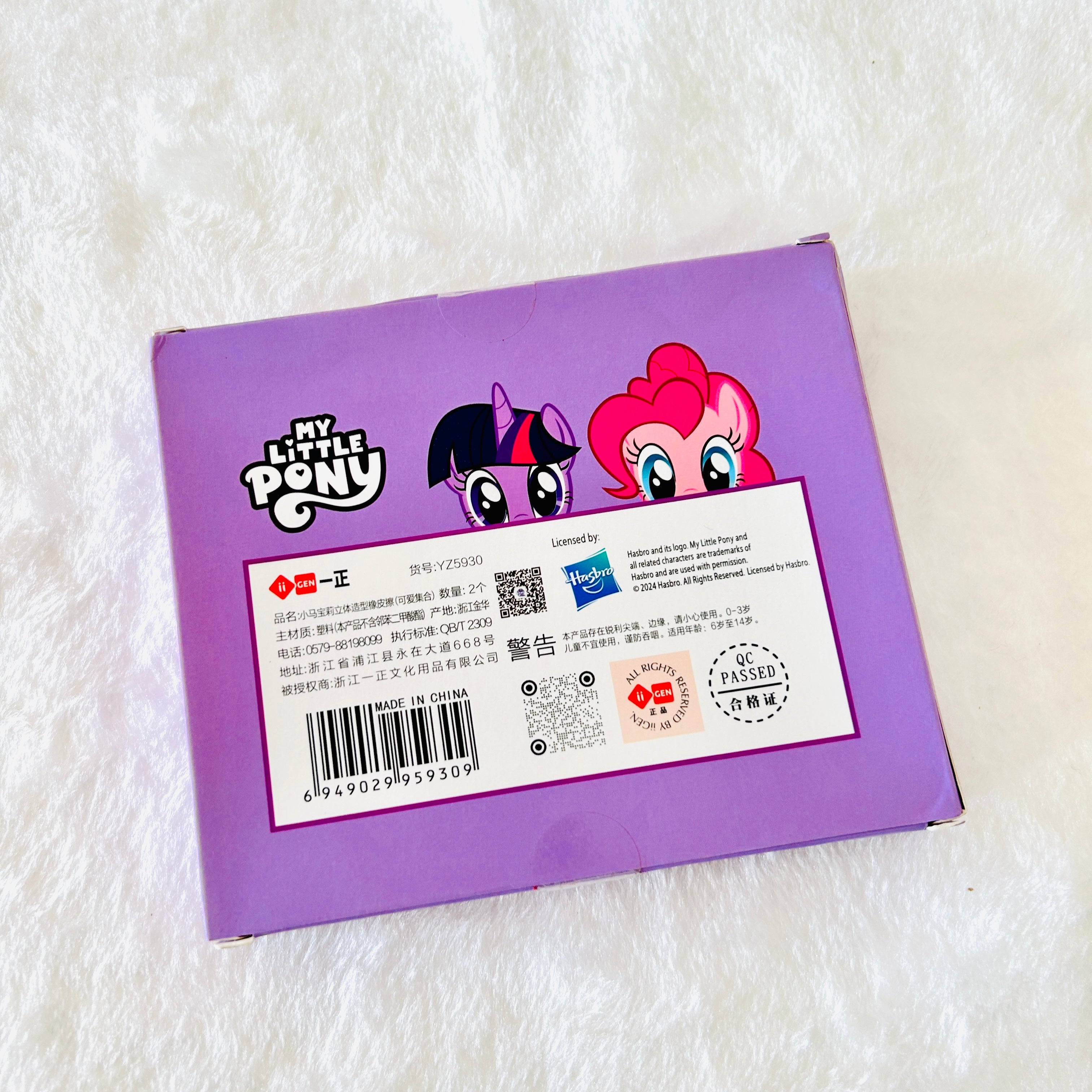 The back of a purple My Little Pony toy box, featuring character artwork of Twilight Sparkle and Pinkie Pie, with product text, a barcode, and a Hasbro logo, photographed on a white textured fabric background.
