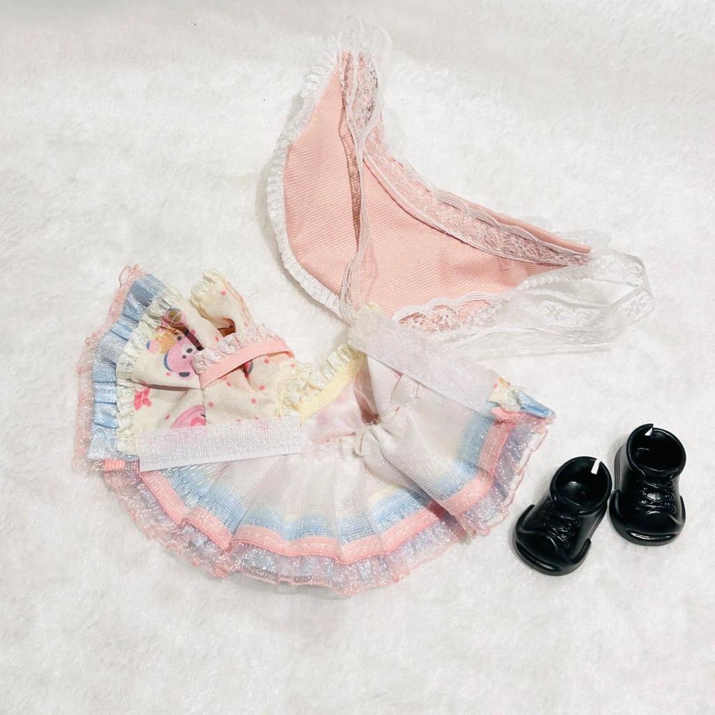 A back view of a doll outfit set featuring a pastel pink and blue ruffled skirt with a floral apron overlay, matching pink lace underwear, and a pair of small black shoes.