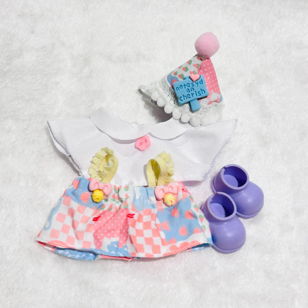 A doll outfit set including a white top, a pink and blue patterned skirt, a matching pink and blue hat with a pom-pom, and a pair of purple booties.