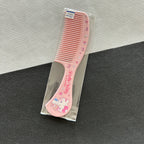 A pink Hello Kitty hair comb with rhinestone accents and a Hello Kitty illustration on the handle, shown inside a clear plastic sleeve on a textured gray and white surface.