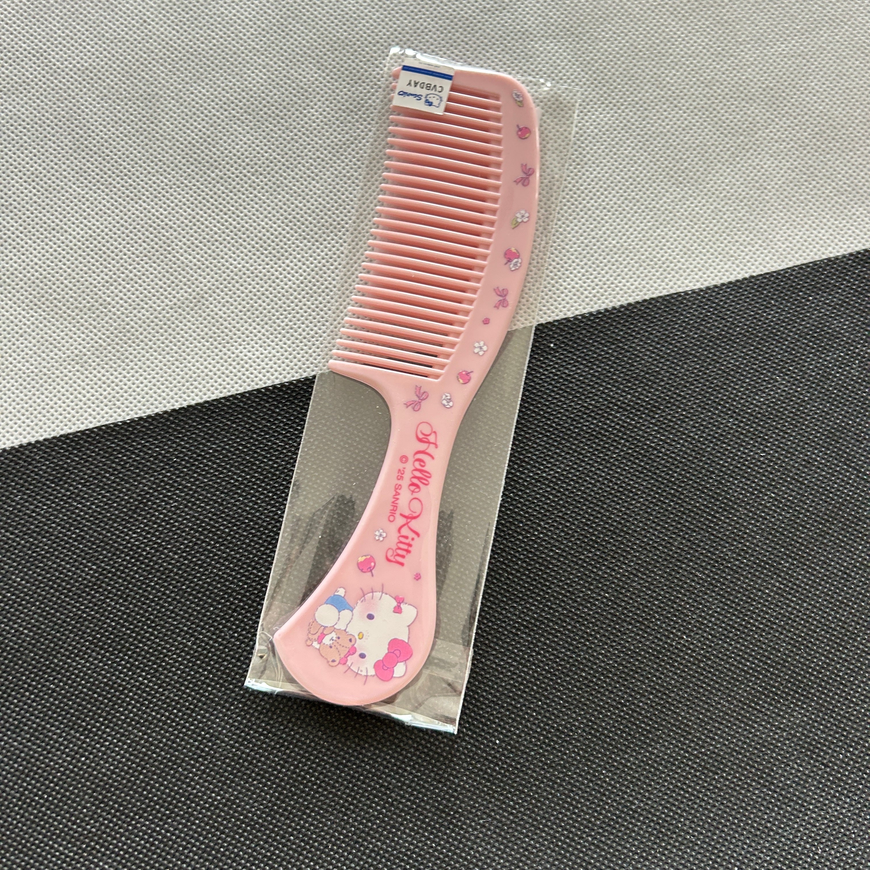 A pink Hello Kitty hair comb with rhinestone accents and a Hello Kitty illustration on the handle, shown inside a clear plastic sleeve on a textured gray and white surface.