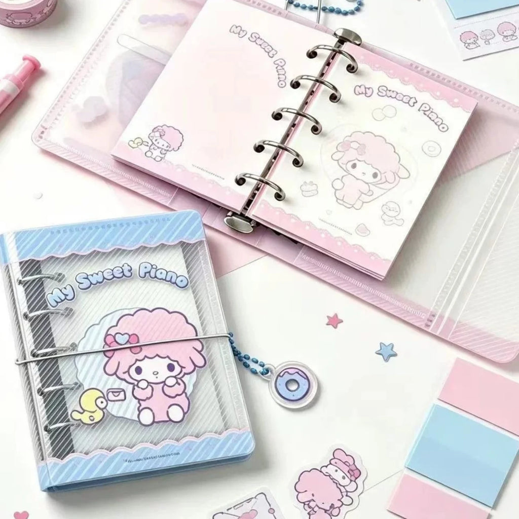 A styled flat-lay of pastel stationery featuring a pink and blue 'My Sweet Piano' ring binder notebook, a matching clear plastic sleeve, stickers, sticky notes, and a small charm on a white background.