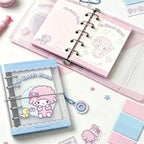 A styled flat-lay of pastel stationery featuring a pink and blue 'My Sweet Piano' ring binder notebook, a matching clear plastic sleeve, stickers, sticky notes, and a small charm on a white background.