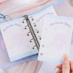 A close-up of a pastel ring binder with cute cartoon notepad pages. A hand is holding one page in front of another that is inserted into the binder.