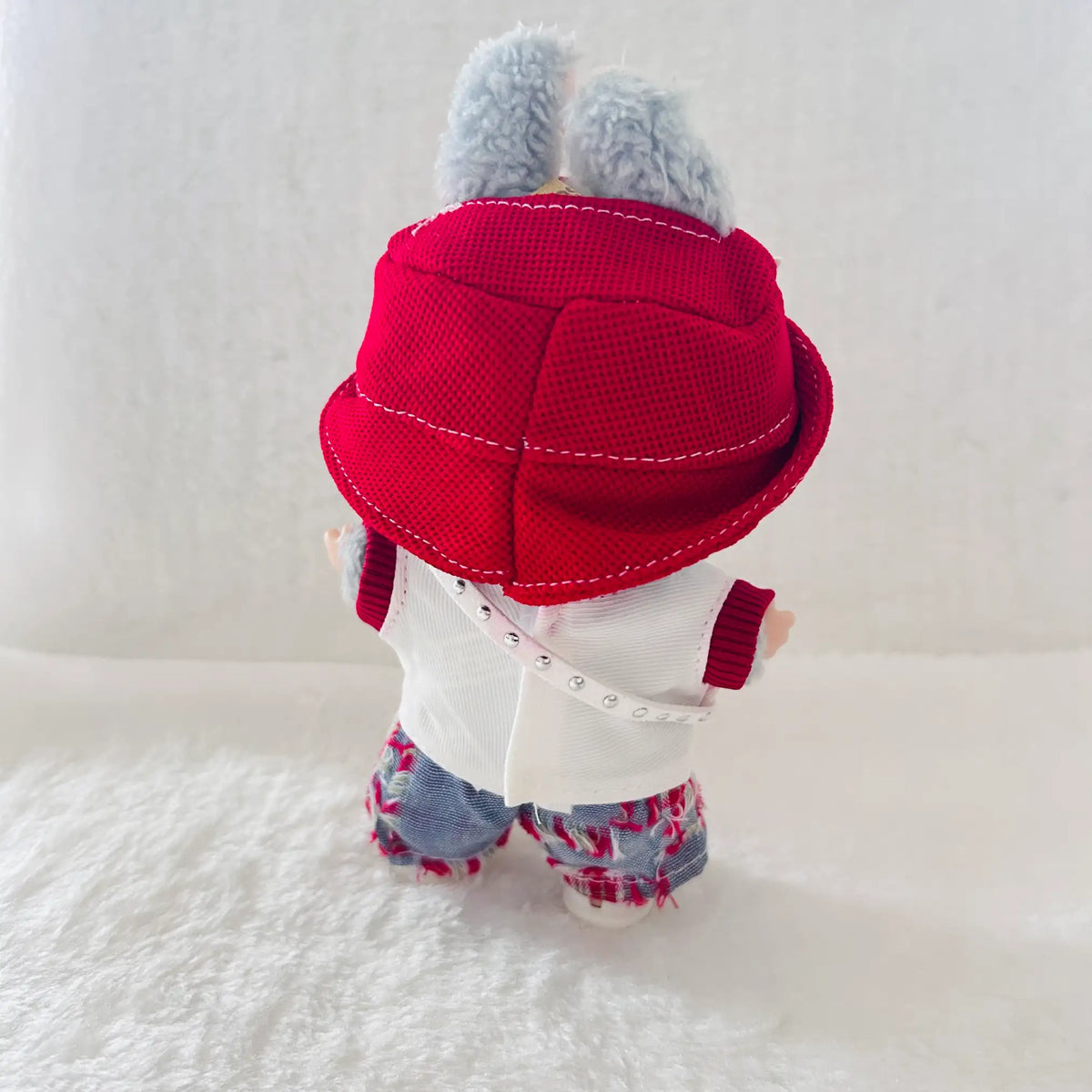 A small plush doll or stuffed animal shown from the back, wearing a bright red bucket hat with gray fuzzy bunny ears, a white top, and blue floral-patterned shorts.