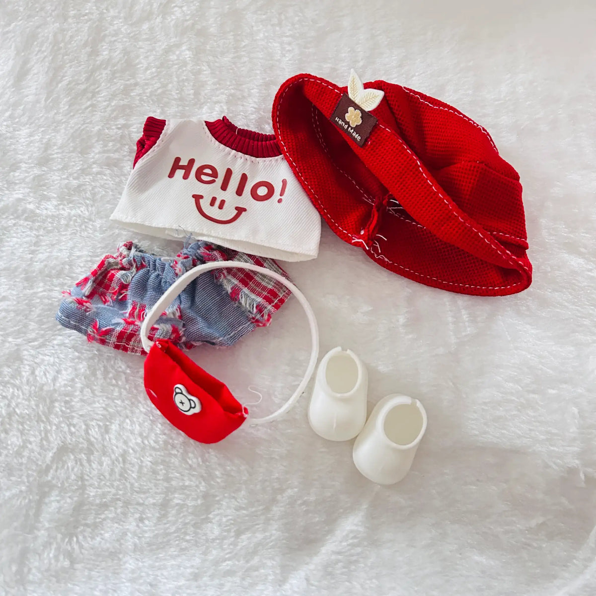 A doll outfit set laid flat on a white textured fabric background, including a red cap, a white top with red 'Hello!' text, red-and-blue plaid shorts, a small red pouch, and a pair of white booties.