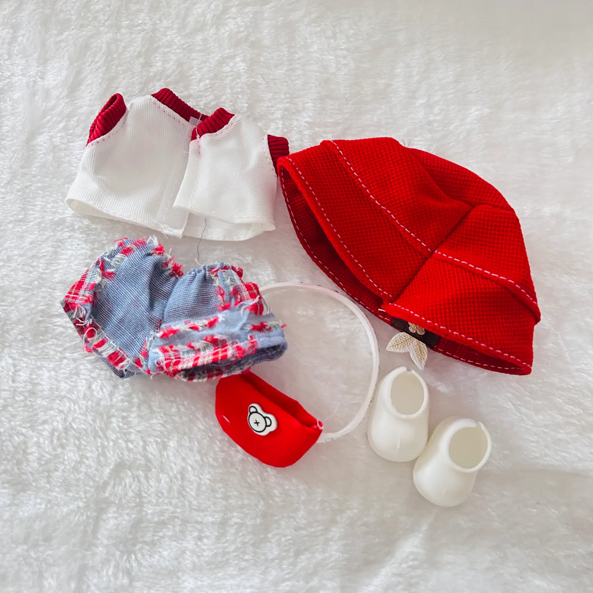 A doll outfit set including a red mesh hat, white top and skirt pieces, a blue-and-red plaid skirt, a small red pouch, and two white bootie-like pieces.