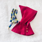 A small red cape accessory with small buttons, laid flat on a white textured background next to a blue, gray, and yellow striped fabric loop.