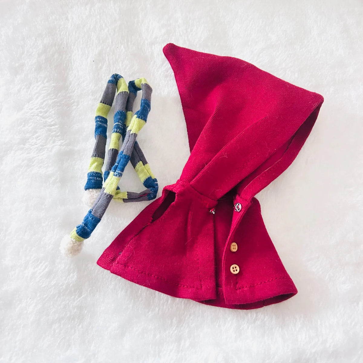 A small red cape accessory with small buttons, laid flat on a white textured background next to a blue, gray, and yellow striped fabric loop.