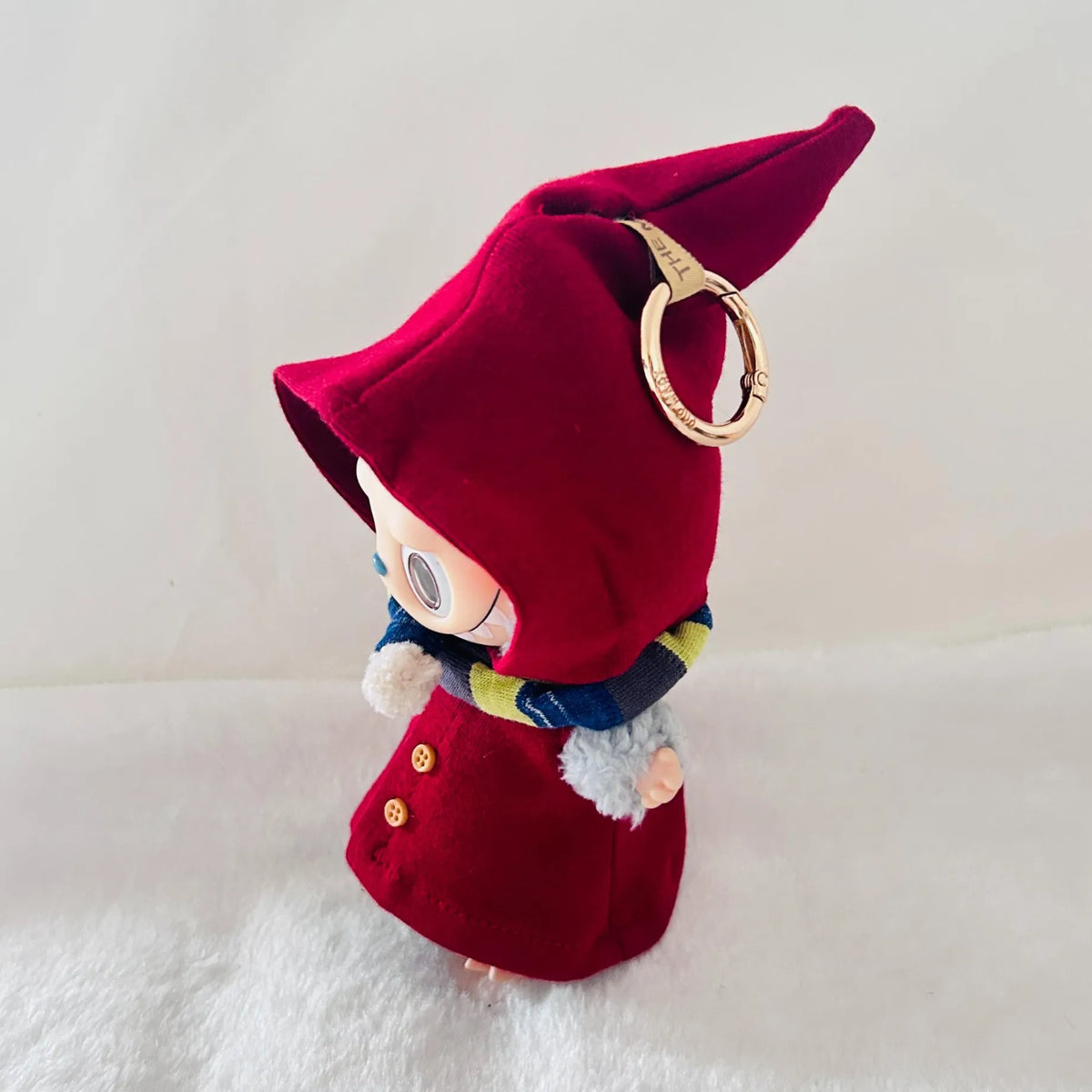 A Labubu plush character doll wearing a deep red pointed hat cape, with a blue and yellow striped scarf.