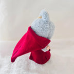 A Labubu plush toy wearing a bright red cape, posed on a soft white textured surface.