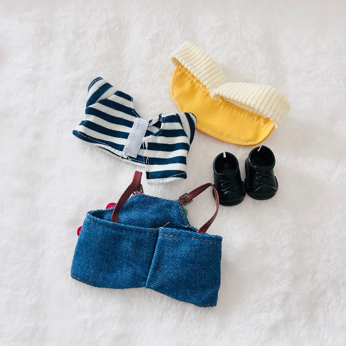 Back view of a set of doll clothing, including a blue denim skirt with brown straps, and a pair of black miniature shoes.