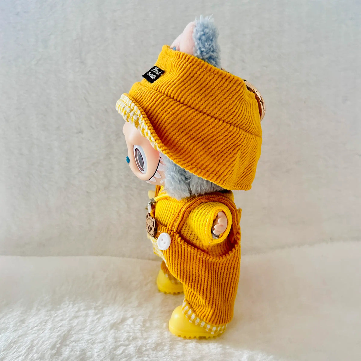 A small plush toy or doll dressed in a mustard yellow corduroy outfit, including a matching hat and yellow booties.