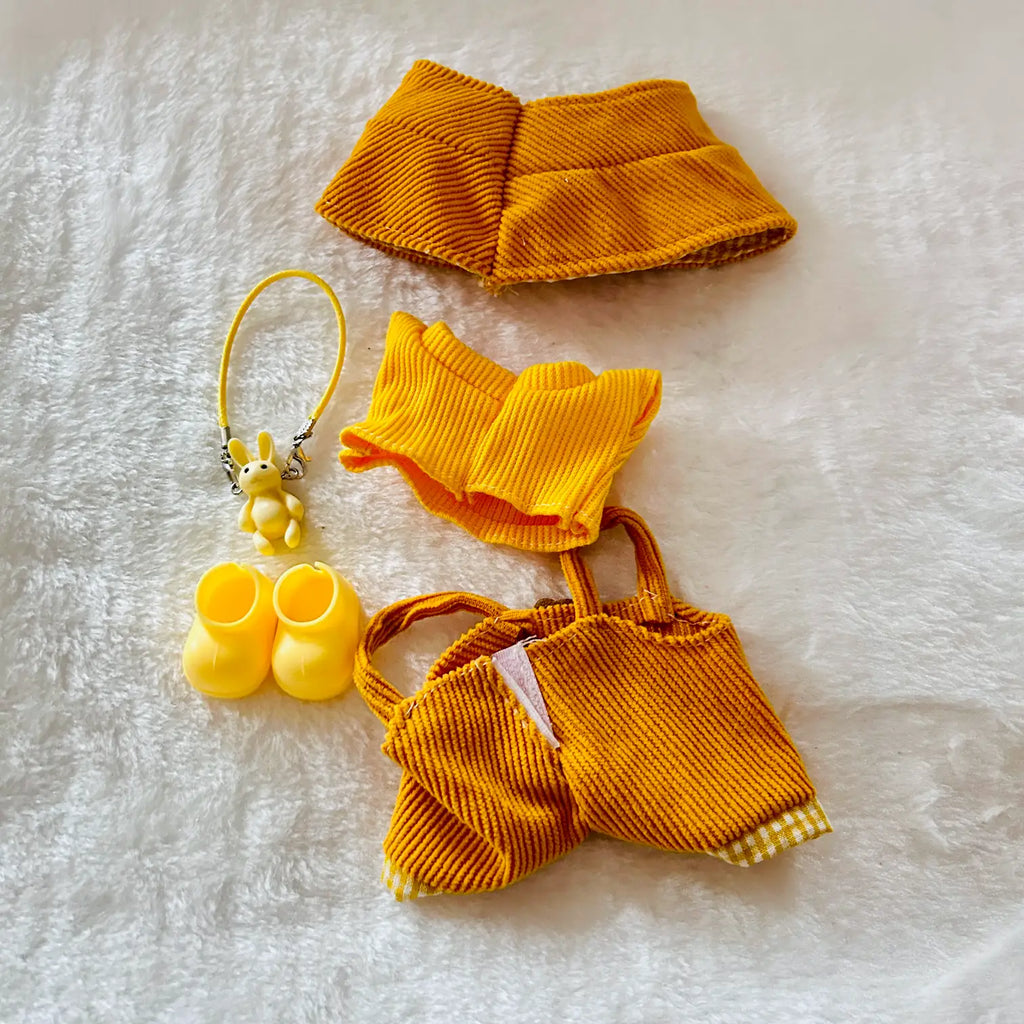 A set of yellow corduroy pet accessories laid out on a white textured blanket, including a collar, a small top, a tote bag, and matching booties with a small plush keychain.