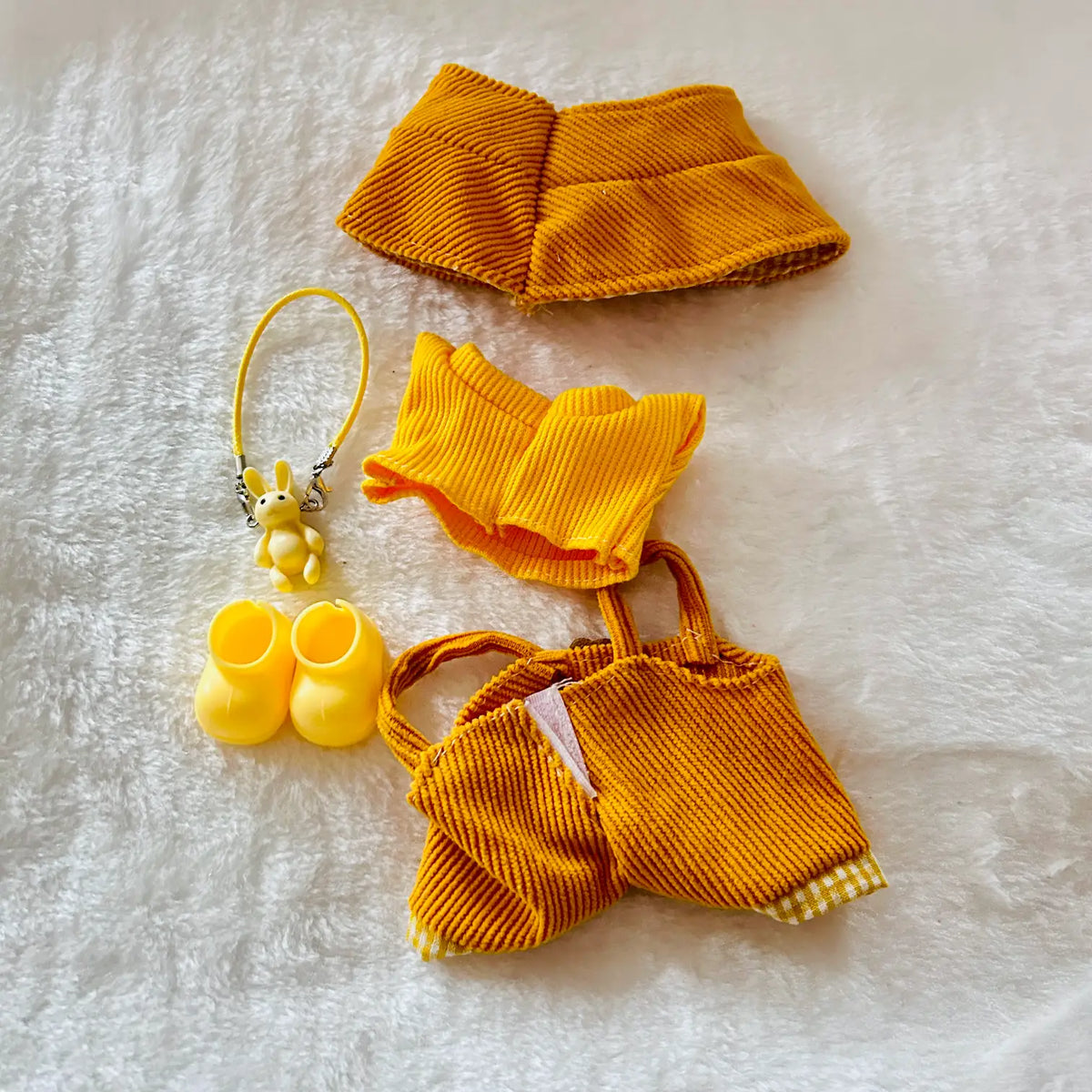 A set of yellow corduroy pet accessories laid out on a white textured blanket, including a collar, a small top, a tote bag, and matching booties with a small plush keychain.