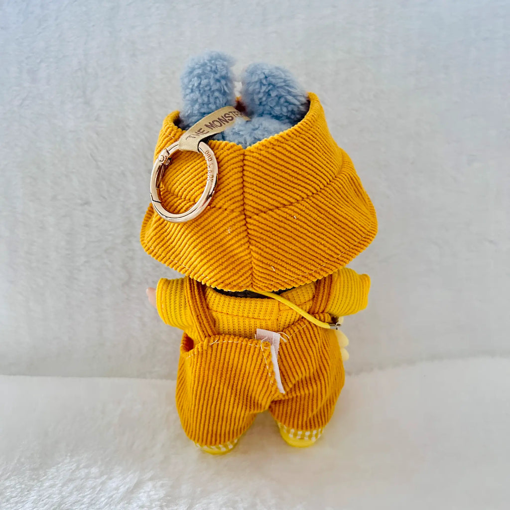 A plush bunny toy shown from the back, wearing a mustard yellow corduroy jumpsuit with blue fluffy ears and a gold metal key ring accessory attached.