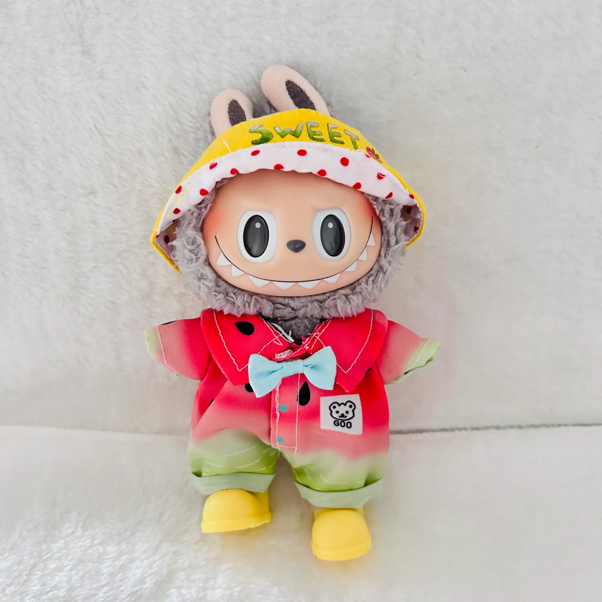A plush toy doll wearing a yellow strawberry hat and a red watermelon-themed outfit with a light blue bow tie.
