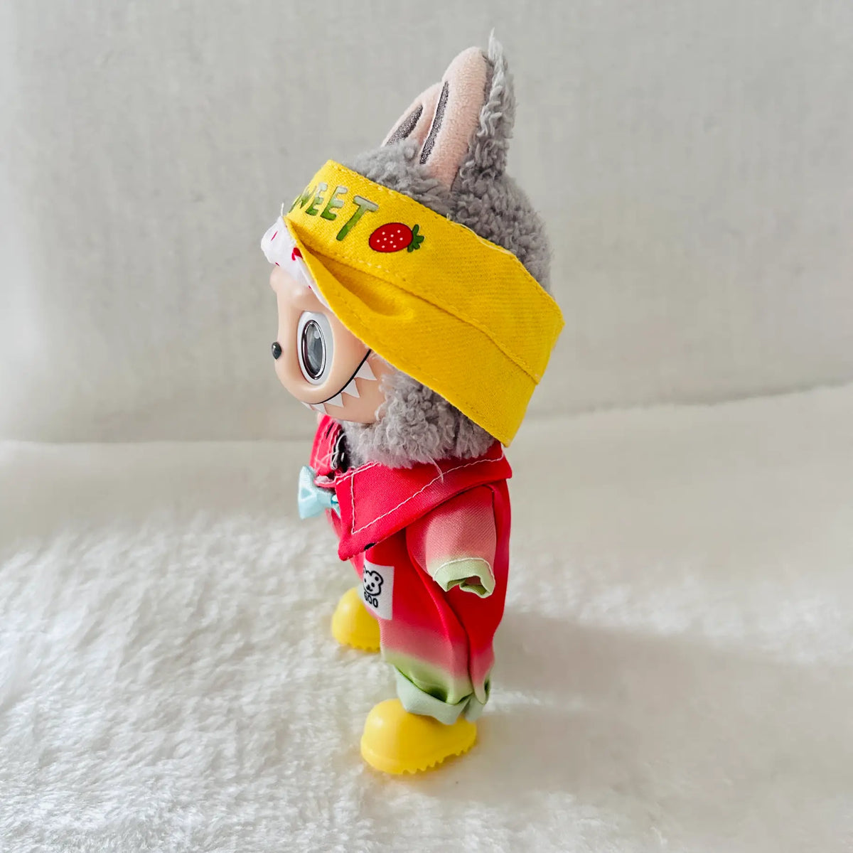 A small plush toy character standing on a soft white surface, wearing a bright yellow headband with green and red lettering and a strawberry patch, plus a red outfit with yellow boots.